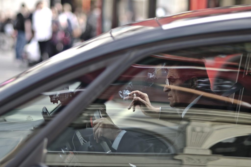 well-dressed man driving a car, viewed from the passenger-side window with reflections layered across the glass.