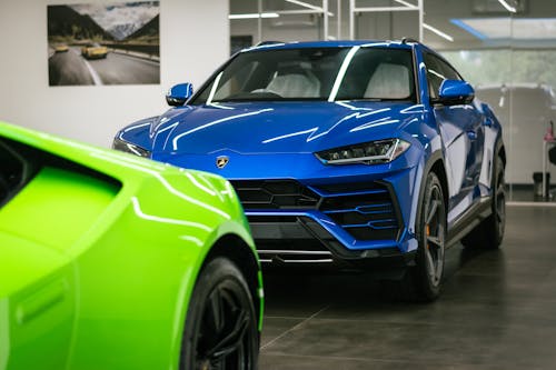 The image shows a vibrant blue Lamborghini Urus SUV prominently displayed in a modern showroom, partially framed by the front of a bright green Lamborghini Huracán in the foreground, highlighting the brand’s bold design and luxury performance appeal.