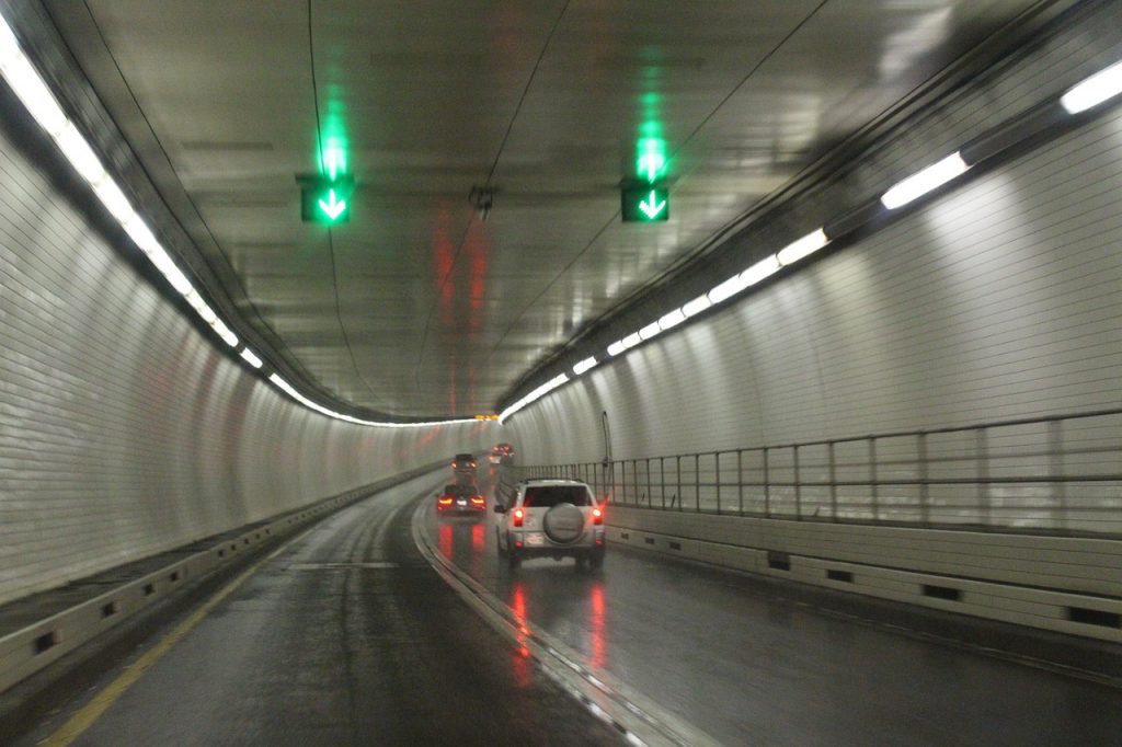 Cars travel through a curved, illuminated tunnel with wet pavement and green lane arrows, creating a sleek and reflective urban driving scene.