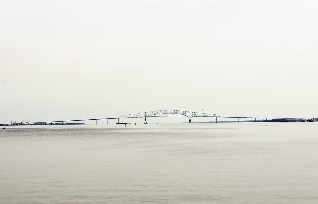 The image shows the Francis Scott Key Bridge in Baltimore, Maryland, stretching across the Patapsco River with its iconic steel arch rising over the water, set against a muted, overcast sky typical of the city’s harbor views.
