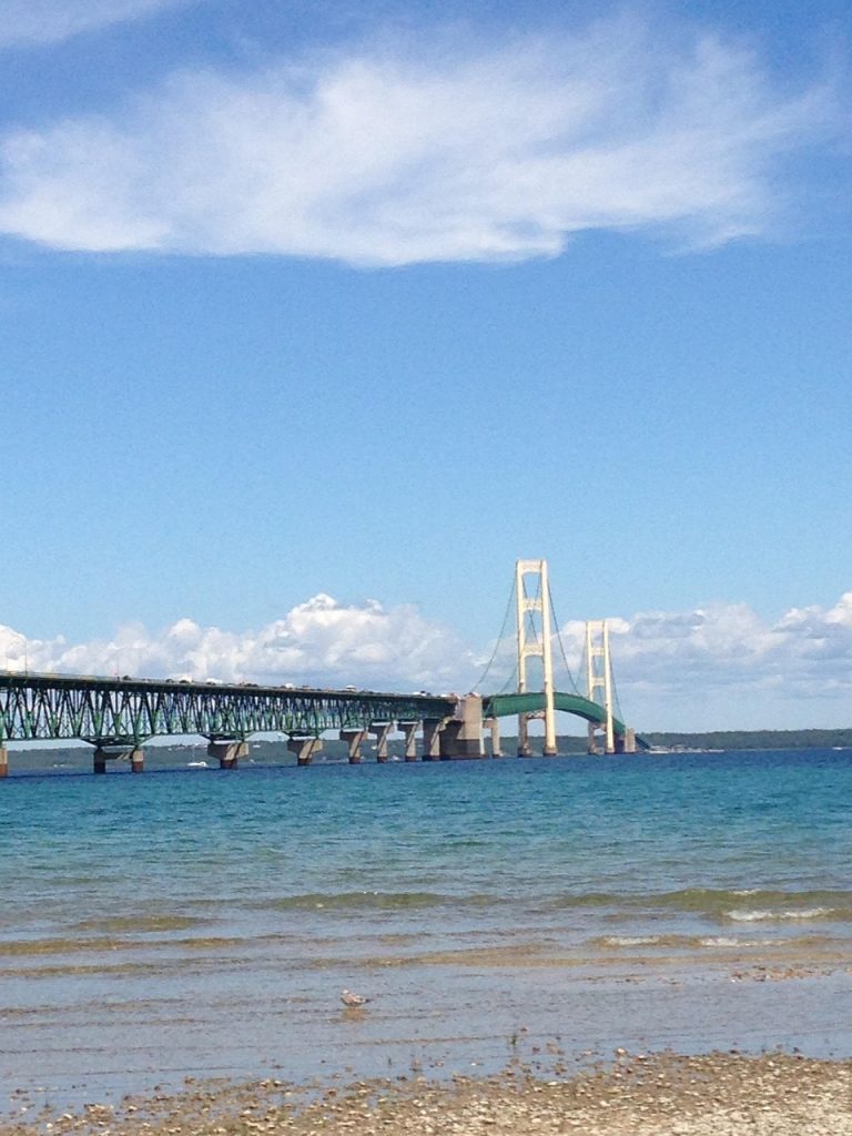 The image shows Michigan’s iconic Mackinac Bridge stretching across the blue waters of the Great Lakes under a bright, partly cloudy sky, connecting Michigan’s Upper and Lower Peninsulas.
