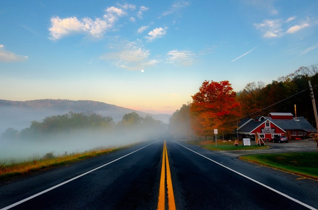 A peaceful Massachusetts road cuts through misty hills at sunrise, flanked by vibrant fall foliage and a classic red country store.