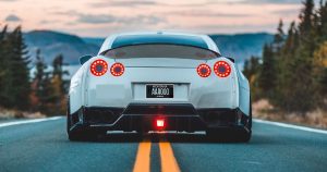 back of a white sports car with a Montana license plate, parked on a two-lane road with a forested landscape on one side and a lake and mountains on the other.