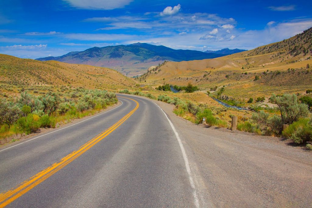 A paved road with a yellow line curves through a dry, mountainous landscape under a bright blue sky with a stream visible in the valley to the right.