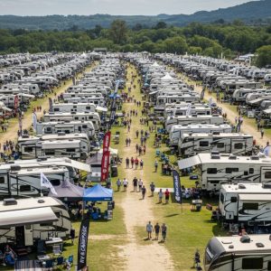 An expansive RV and Camper Showcase is underway in an open field, where numerous RVs of all sizes are parked in orderly rows, with people exploring the vehicles and browsing vendor tents under a partly cloudy sky.
