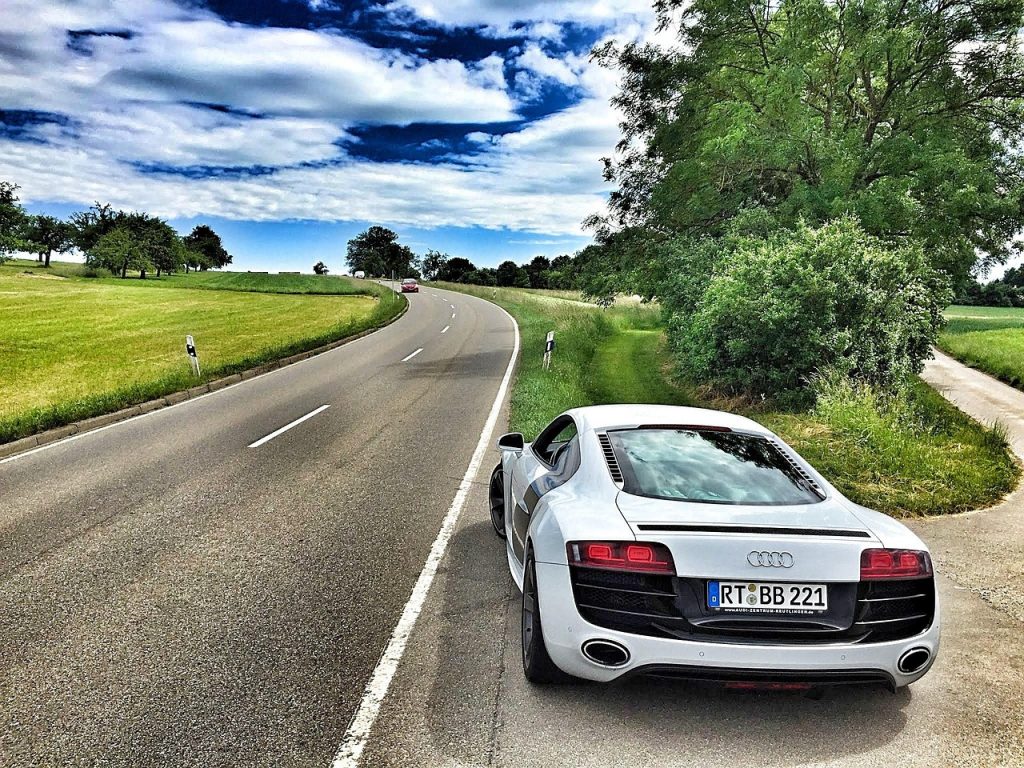 A sleek white sports car is parked along a winding country road surrounded by green fields and trees under a bright, partly cloudy sky.