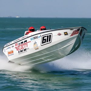 White Velocity offshore racing boat (Team Ride Legal, number 611) with two crew members, slightly airborne on choppy blue water during a race.