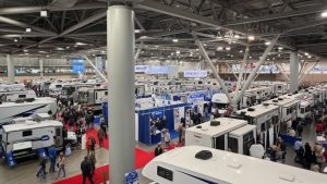 Overhead view of a crowded indoor RV show featuring a wide variety of motorhomes and travel trailers with attendees walking on red carpet aisles.